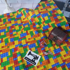 Puede incluir: Una tela colorida con un patrón repetitivo de bloques de construcción de juguetes en rojo, azul, verde, amarillo y naranja. La tela tiene una superficie texturizada y es perfecta para proyectos de manualidades.