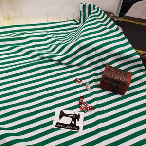 Puede incluir: Tela a rayas verdes y blancas con un pequeño cofre de madera y pinzas de costura. También es visible una pegatina con una máquina de coser y el texto "RUBYJAM FABRIC". La tela está doblada y drapeada.
