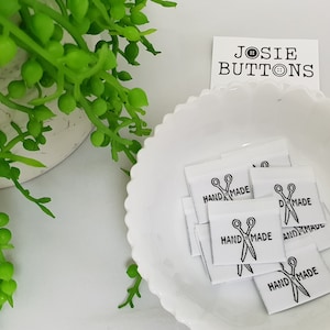 Puede incluir: Un bol blanco lleno de etiquetas de tela blanca con texto negro y un gráfico de tijeras. Las etiquetas dicen "HANDMADE". Una tarjeta blanca con texto negro dice "JOSIE BUTTONS".