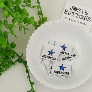 Puede incluir: Un bol blanco lleno de 10 etiquetas tejidas blancas con una estrella azul y el texto "SUPERSTAR" impreso en ellas. Las etiquetas son para ropa y se venden por Josie Buttons.