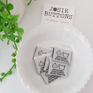 Puede incluir: Un bol blanco con 10 etiquetas tejidas blancas que dicen "Quitar el bebé de la prenda antes de lavar" en texto negro. Las etiquetas están en un bol blanco sobre una superficie blanca. El texto "JOSIE BUTTONS 10 Woven Labels" está en una tarjeta blanca en el fondo.