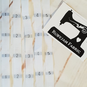 Puede incluir: Etiquetas de tela blancas con números grises del 1 al 5. Las etiquetas están dispuestas verticalmente. Un logotipo de Rubyjam Fabric en blanco y negro está en la esquina superior derecha. Para coser y manualidades.