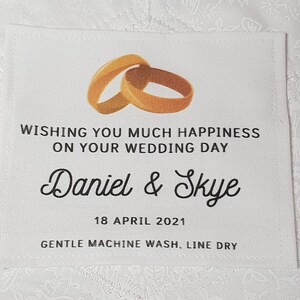 Puede incluir: Etiqueta de tela blanca con anillos de boda dorados, el texto "WISHING YOU MUCH HAPPINESS ON YOUR WEDDING DAY", los nombres "Daniel & Skye", la fecha "18 APRIL 2021" y las instrucciones "GENTLE MACHINE WASH, LINE DRY".