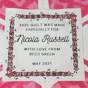 May include: A white fabric label with a pink heart border. The label reads "This quilt was made especially for Nicola Russell with love from Becs Green May 2021".