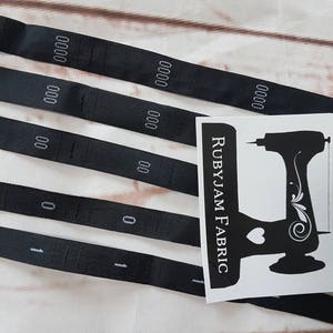 Puede incluir: Cinco tiras de tela negra con números blancos impresos y costuras. Las tiras están dispuestas en fila, siendo la tira superior la más larga y la tira inferior la más corta. Una pegatina blanca con el texto "RUBYJAM FABRIC" y una silueta negra de una máquina de coser se encuentra en la esquina superior derecha de la imagen.