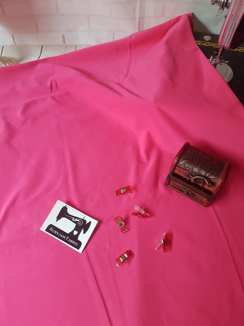 May include: A bright pink fabric swatch with a small white label that says "RubyJam Fabric" and a sewing machine icon. There are also several red plastic clips scattered on the fabric.
