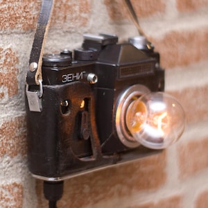 May include: A black vintage Zenit camera repurposed as a wall lamp, featuring a clear spherical light bulb protruding from the lens mount. The camera body is encased in a dark brown leather case with a matching strap and displays the Cyrillic text "ЗЕНИТ".