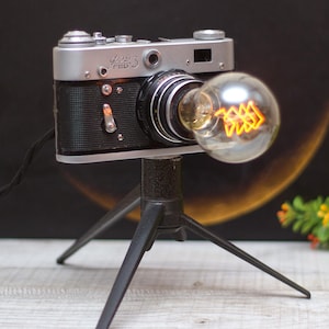 May include: A vintage camera lamp with a silver and black camera body, a clear glass light bulb, and a black tripod stand.