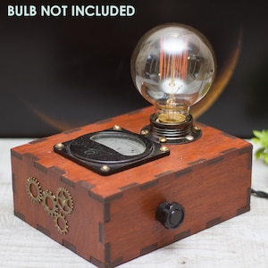 Könnte beinhalten: Eine Steampunk-Tischlampe aus Holz mit einer klaren Glühbirne aus Glas, einem schwarzen Zifferblatt und einem Knopf an der Seite. Die Lampe ist aus Holz und hat eine braune Oberfläche. Der Text "BULB NOT INCLUDED" ist im Bild sichtbar.