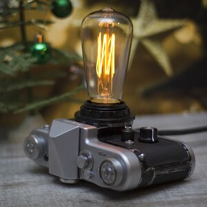 May include: A vintage silver and black film camera repurposed as a table lamp. The camera body features metallic dials and a textured grip, topped with a clear glass Edison-style light bulb that emits a warm, glowing orange filament light.
