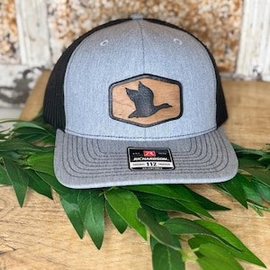 May include: Gray and black trucker hat with a duck emblem. The hat features a gray front panel with a black mesh back and a curved brim. A leather patch with a duck silhouette is centered on the front. The hat has the text "RICHARDSON ORIGINAL 112 TRUCKER ADJUSTABLE".
