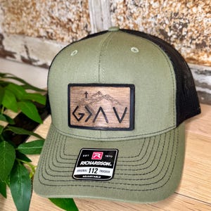 May include: A green and black trucker hat with a wood-look patch that says "G>A<V" with a cross above the "G". The hat is adjustable and has the Richardson logo on the back.