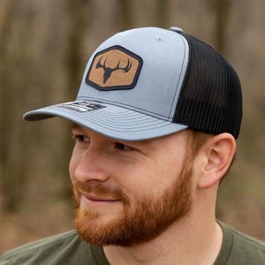 May include: A light blue and black trucker hat with a brown leather patch featuring a deer antler design. The hat has a curved brim and a mesh back for breathability. A stylish accessory for outdoor enthusiasts.