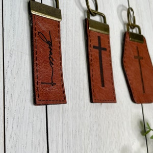 May include: Three leather keychains with metal rings. The first keychain is brown and has the word "Grace" engraved on it with a small cross below. The second keychain is brown and has a cross engraved on it. The third keychain is brown and has a cross engraved on it in a different shape.