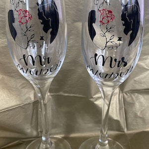 May include: Two clear glass champagne flutes with black and red designs. The flutes are decorated with a silhouette of Belle and the Beast from the movie Beauty and the Beast. The flutes are labelled "Mr" and "Mrs" with the date "04/07/2022".