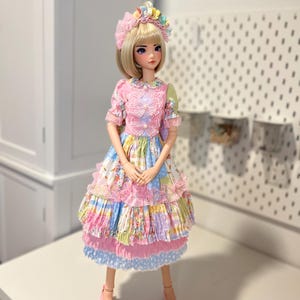 May include: A doll wearing a pink, blue, and yellow dress with a floral pattern and a white lace trim. The doll has blonde hair and blue eyes and is wearing a pink flower crown.