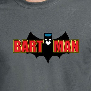 May include: Dark gray t-shirt featuring a Bart Simpson-inspired design. The graphic includes a black bat symbol with Bart's face and the words "BART MAN" in red and yellow lettering. The shirt is a short-sleeved crew neck.