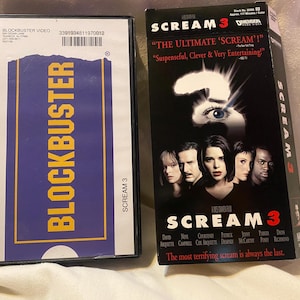 Peut inclure: Un boîtier VHS de location Blockbuster Video avec une étiquette bleue et jaune à côté d'une boîte de film VHS Scream 3 avec une couverture noire, un masque de fantôme blanc, le titre en rouge et une photo des acteurs avec le texte The Ultimate Scream.