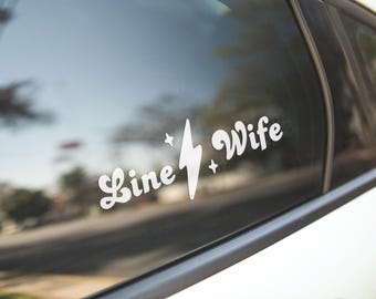 May include: White vinyl decal with the text "Line Wife" and a lightning bolt graphic. The decal is on a car window.