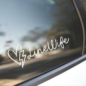 May include: White vinyl decal applied to a car window. The decal features a heart, a lightning bolt, and the script text "Linell Wife". The car window reflects the outside environment.