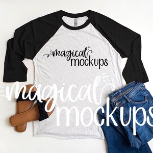 May include: A white and black baseball tee shirt with long sleeves. The shirt has the text "magical mockups" printed on the front. The shirt is laid flat on a white surface with a pair of brown sandals and blue jeans.
