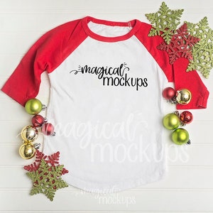 May include: A white long-sleeved t-shirt with red sleeves, a white background, and the text "magical mockups" printed on the front. The shirt is surrounded by red and green Christmas ornaments and snowflakes.