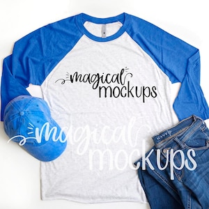 May include: A white and blue baseball tee shirt with the text "magical mockups" printed on the front. The shirt is paired with a blue baseball cap and blue jeans.