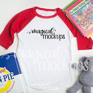 May include: A white and red baseball tee shirt with the text "magical mockups" printed on the front. The shirt is lying on a white wooden surface with a blue and yellow box, a gray stuffed elephant, and a stack of children's books.