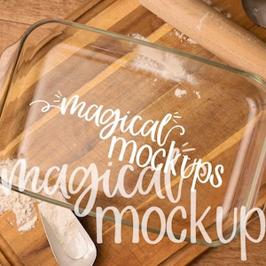 May include: A clear glass baking dish on a wooden cutting board with flour scattered around it. The dish is empty and ready to be used for baking. The text "magical mockups" is written on the dish in white.
