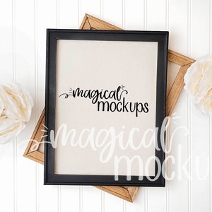 May include: A black frame with a white canvas insert, featuring the text "magical mockups" in black script. The frame is on a white wood background with two white flowers.