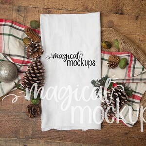 May include: White kitchen towel with the text "magical mockups" written in black cursive. The towel is surrounded by pine cones, burlap, and a red and white plaid kitchen towel.