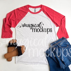 May include: A white and red baseball tee shirt with the text "magical mockups" printed on the front. The shirt is laid flat on a white surface with a pair of brown sandals and blue jeans.