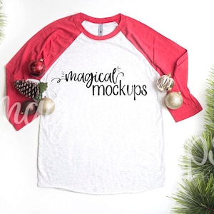 May include: A white and red baseball tee shirt with the text "magical mockups" printed on the front. The shirt is laying flat on a white surface with pine branches and red and gold ornaments around it.