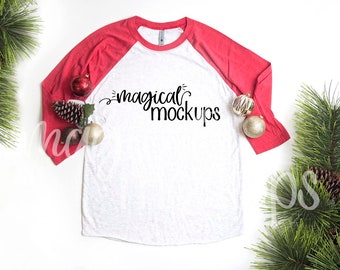 Puede incluir: Una camiseta de béisbol blanca y roja con el texto "magical mockups" impreso en la parte delantera. La camiseta está colocada sobre una superficie blanca con ramas de pino y adornos rojos y dorados alrededor.