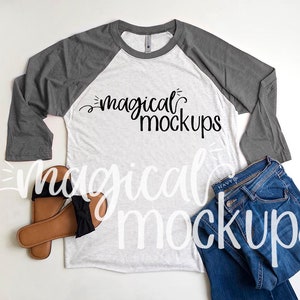 May include: A white and gray long-sleeved t-shirt with a raglan design. The shirt has the text "magical mockups" printed on the front. The shirt is laid flat on a white surface with a pair of brown sandals and blue jeans.