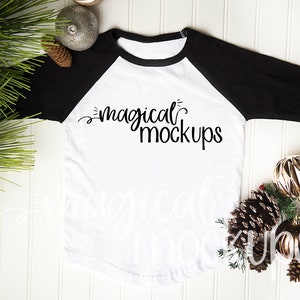 May include: White and black baseball tee shirt with the text "magical mockups" printed on the front. The shirt is laying flat on a white wood surface with pine branches and gold ornaments.