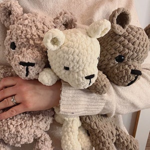 May include: Three crocheted teddy bears in different colors: beige, white, and brown. The bears are held in the arms of a person.