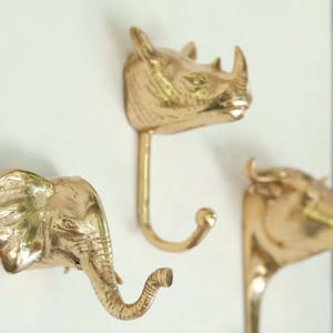 Könnte beinhalten: Drei Wandhaken aus Messing in Form von Tierköpfen: ein Elefant, ein Nashorn und ein Bulle. Die Haken sind goldfarben und an einer weißen Wand befestigt. Der Elefantenhaken hat einen gebogenen Rüssel.