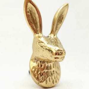 Pode incluir: Estatueta de coelho dourado com orelhas longas e corpo texturizado. O coelho é um item decorativo, possivelmente um puxador de gaveta ou puxador de armário, em fundo branco.