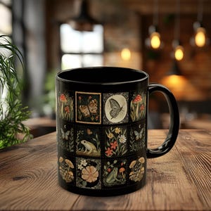May include: Black ceramic mug featuring a collage of vintage-style illustrations. The design includes butterflies, mushrooms, flowers, and a frog. The mug has a black handle and a glossy finish, perfect for hot beverages.