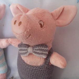 Può includere: Un maiale di peluche rosa con una salopette di maglia grigia e un papillon a righe bianche e grigie.
