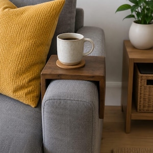 May include: A rectangular wooden armrest tray with a dark brown finish, designed to sit over the arm of a sofa. A round cork coaster is placed on the centre of the tray, supporting a speckled off-white ceramic mug filled with a dark liquid.