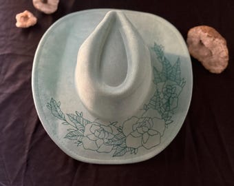 May include: A light blue cowboy hat with a green floral design. The hat has a wide brim and a tall crown.