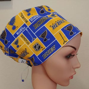 May include: A blue and gold St. Louis Blues scrub cap. The cap features a repeating pattern of the team's logo, hockey sticks, and the words "Blues" and "St. Louis". The cap has a white drawstring for adjustment.