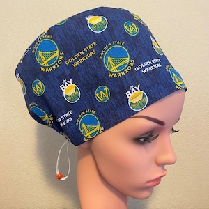 Peut inclure: Un bonnet de chirurgien bleu et jaune avec le logo des Golden State Warriors répété sur tout le tissu. Le bonnet a un cordon de serrage blanc.