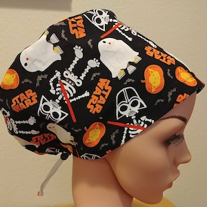 May include: Black scrub hat with a Halloween Star Wars theme. The hat features a repeating pattern of skeletons, pumpkins, bats, and Star Wars characters like Darth Vader and Yoda. The text "Star Wars" is repeated throughout the pattern.