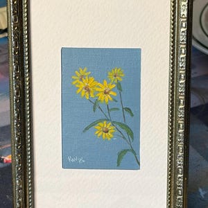 Puede incluir: Una pequeña pintura rectangular de flores amarillas con tallos y hojas verdes sobre un fondo azul claro, dentro de un marco metálico dorado ornamentado. La obra presenta una firma en la esquina inferior izquierda que dice "Kav 26".