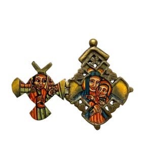 Pode incluir: Um pingente de latão articulado com duas seções em forma de cruz com figuras religiosas pintadas. A seção esquerda retrata uma figura em laranja e verde, enquanto a direita mostra duas figuras com vestes azuis e laranjas sobre fundo amarelo.