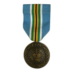 Op de afbeelding: Een bronzen VN-medaille met een lichtblauw lint. De medaille toont het VN-embleem en de letters "UN". Het lint heeft strepen in wit, groen, geel en marineblauw.
