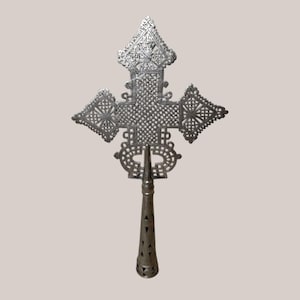 May include: Ornate silver-colored cross with intricate geometric patterns. The cross features a detailed design with a central diamond-shaped element and decorative edges. The handle is conical with triangular cutouts, adding to the cross's unique aesthetic.